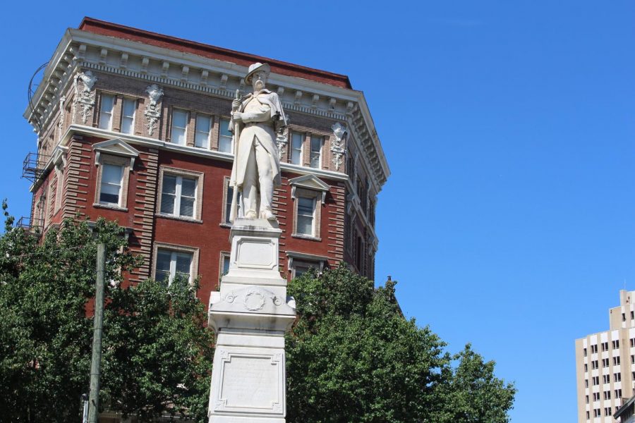 An image of Macons Confederate Monument. He is a man made of white marble standing on a tall white marble platform and holding a rifle. Behind him is a tall brown buildin, green trees and a clear blue sky.