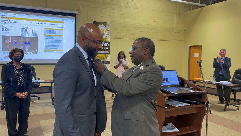 Superintendent Curtis Jones, left, secures a Victory in Progress pin to incoming Superintendent Dan Sims' coat at the May 10 meeting solidifying Sims as the district's next leader.